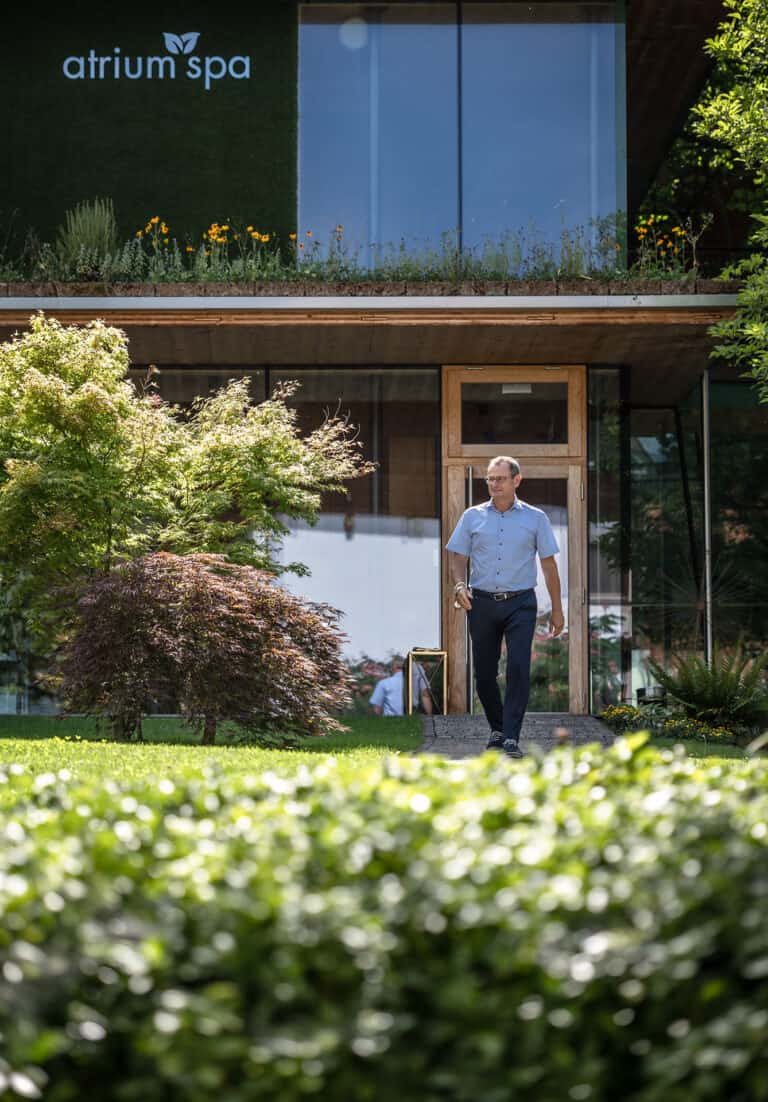 Mann geht durch modernen Garten mit üppigen Pflanzen und Blick auf Atrium Spa am Gebäude.