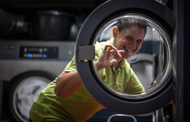 Aliasing: Frau in grüner Arbeitskleidung wäscht im Waschsalon, lacht freundlich in die Kamera.