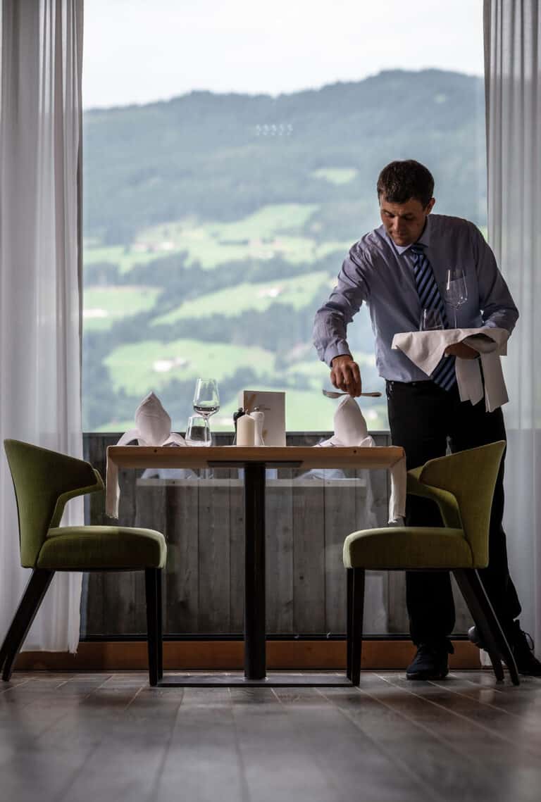 Ein Kellner bereitet einen Tisch im Restaurant vor, mit Blick auf die grüne, bergige Landschaft im Hintergrund.
