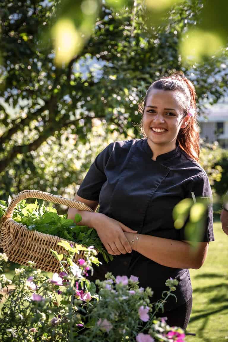 Alt: Junge Gärtnerin hält Korb voller frischer Kräuter im sonnigen Garten.