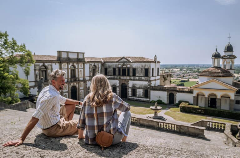 Zwei Personen auf einer Terrasse vor einer historischen Kunst- und Architekturikone in Italien, mit Blick auf alte Gebäude und die Landschaft.