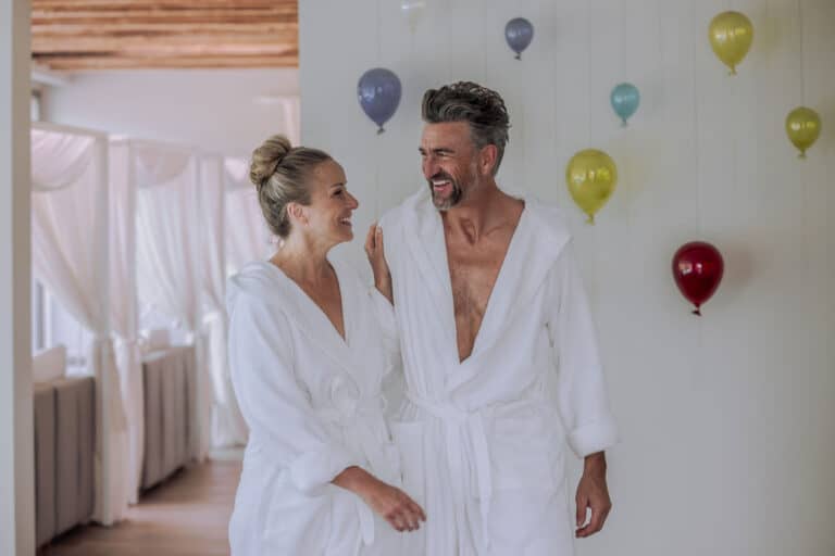 Entspannte Menschen im Bademantel lachen und genießen einen Wellness-Tag, umgeben von Ballons, glückliche Stimmung.