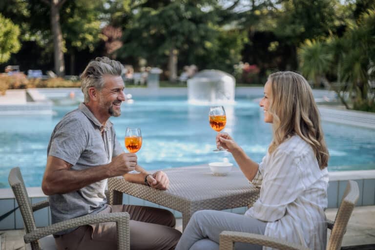 Fröhliches Paar mit Aperol Spritz am Pool, entspannt im Garten, schöne Sonnenuntergang-Atmosphäre.